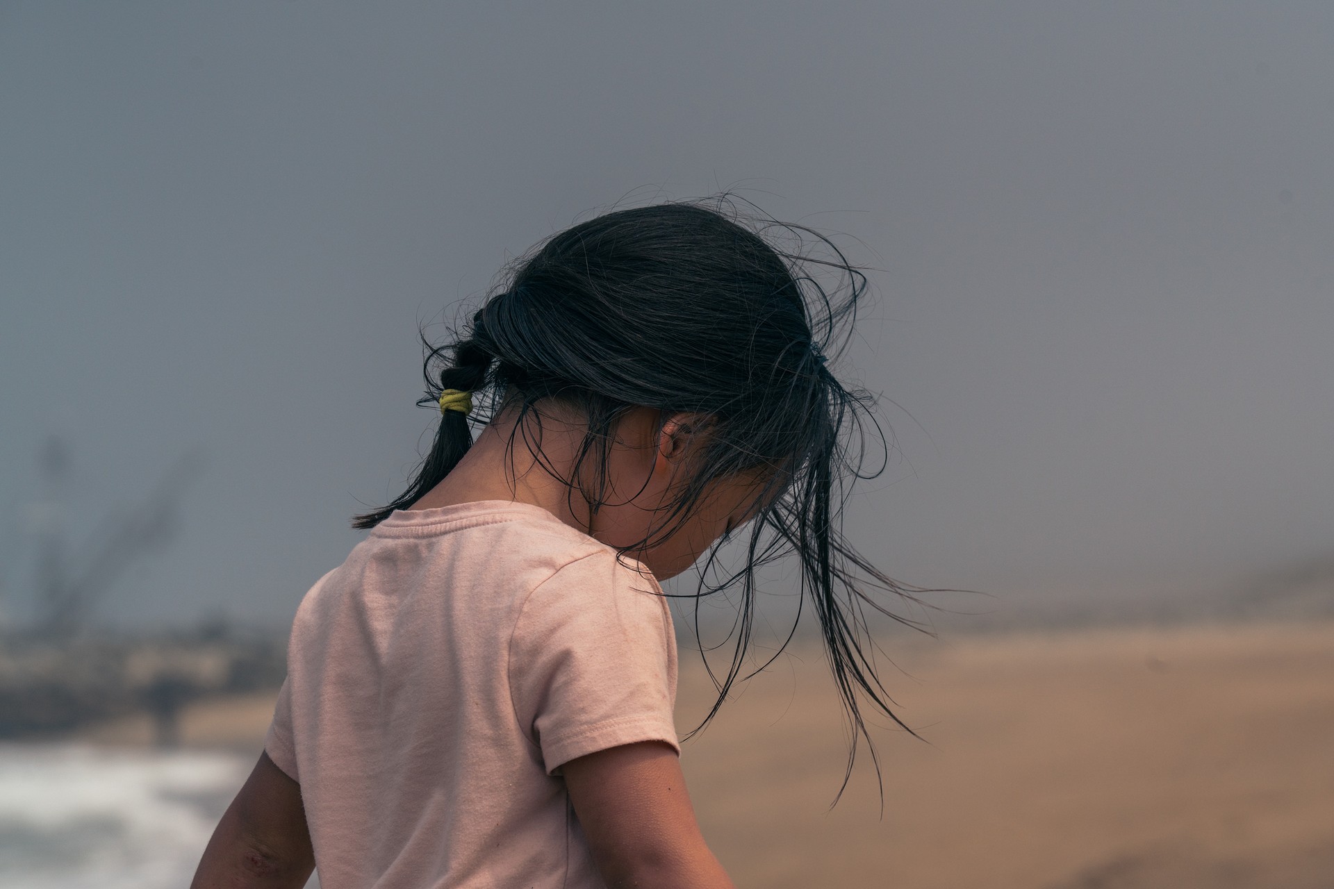 Child at the beach with windblown hair, looking down, overcast sky